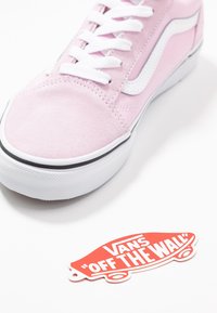 Baskets Vans roses avec lacets blancs et bande latérale à côté d'une étiquette rouge en forme de skateboard "Vans Off The Wall" sur une surface blanche.