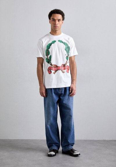 White cotton t-shirt featuring green wreath and red ribbon graphic with "Moschino" text, paired with loose blue denim jeans and black shoes.