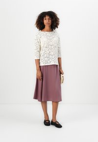 Un haut en dentelle blanc à manches trois-quarts, associé à une jupe midi mauve. Des chaussures noires perforées et une pochette en osier complètent le look.