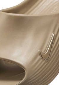 Sandalia deslizante de goma beige con una amplia abertura, superficie texturizada y un prominente logo Nike swoosh en relieve en el lateral.