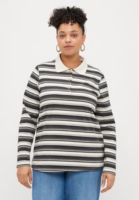 Sieviete ar lockiem, kurai ir zelta auskari-gnome, melnbalts svītrainais krekls ar garām piedurknēm un zili džinsi, skatās uz priekšu.