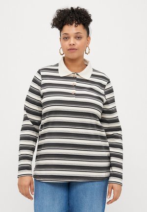 Mulher com cabelo encaracolado usando brincos de argola dourados, uma camisa polo de mangas longas com riscas em preto e branco, e calças jeans azuis, virada para a frente.