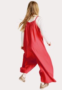 Tuta di cotone rossa con vestibilità ampia, gambe larghe e bretelle elastiche. Abbinata a scarpe marroni e calze bianche a righe.