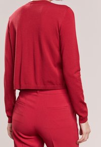 Punainen lyhyt villapaita pitkillä hihoilla, ribbireunalla ja pehmeällä tekstuurilla, yhdistettynä saman värisiin kapeisiin punaisiin housuihin, joissa on takataskut.