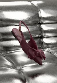 Bordó slingback lapos cipő hegyes orral és nagy masni díszítéssel. Simak felület, enyhe fényességgel, fényes ezüst háttér előtt.