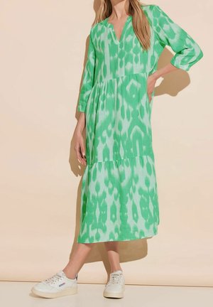 Kvinde iført en grøn mønstret midikjole med 3/4-ærmer og hvide sneakers, stående foran en beige baggrund.