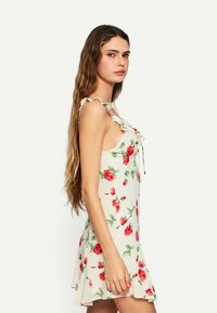Rochie florală cu fundal crem, decorată cu trandafiri roșii, bretele franjurate și o talie strânsă cu detalii de legare. Poale evazate.