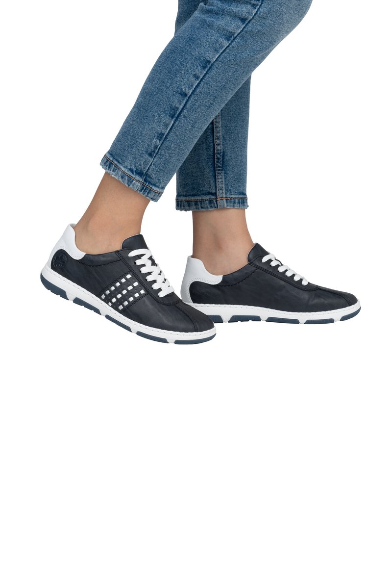 Baskets en cuir noir avec accents blancs, présentant un laçage, des panneaux latéraux texturés et des semelles en caoutchouc, exposées sur un pied vêtu de denim.