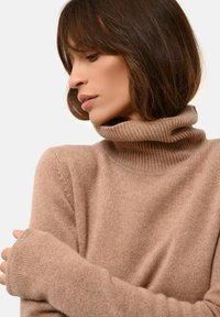Pull à col roulé beige avec des accents en côtes, texture douce et manches allongées. Le tissu semble léger et confortable.