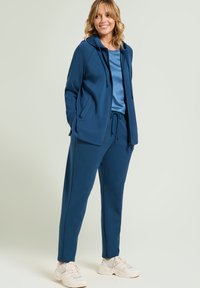 Tiegraues Sportbekleidungsset mit einer Kapuzenjacke mit Taschen und einem Kordelzugbund, kombiniert mit passenden Jogginghosen und cremefarbenen Sneakers.