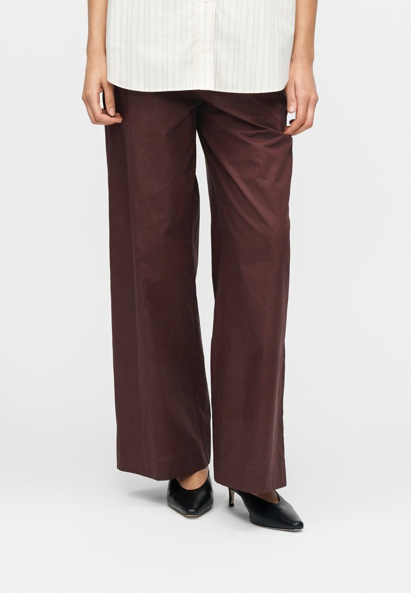 Pantalon à jambes larges de couleur bordeaux riche, en tissu lisse, associé à des chaussures à talons noires. Chemise rayée de couleur claire visible au-dessus.