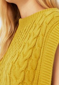 Pull jaune tricoté avec un motif torsadé, un col montant côtelé et un design sans manches en tissu texturé.