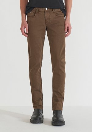 Pantalones de mezclilla marrones con un ajuste slim, bolsillos frontales y cierre de botón. Se llevan con botas negras hasta el tobillo, contra un fondo neutro.