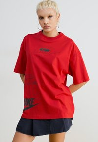 Raudona per didelė medvilninė marškinė su juodu Nike logotipu ir tekstiniais grafikas. Trumpomis rankovėmis ir apvaliais iškirpimais. Lygi tekstūra, kasdienis kirpimas.
