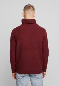 Bordeauxroter Pullover mit hohem Kragen, gerippten Bündchen und strukturiertem Strickmuster. Die Rückseite zeigt ein dezentes Muster und ist tailliert geschnitten.