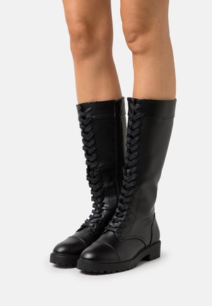 Nogi w czarnych skórzanych sznurowanych butach za kolano na grubych podeszwach, stojące na jasnym, jednolitym tle.