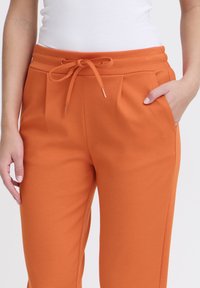 Pantalones de chándal naranjas con cintura ajustable, que cuentan con bolsillos laterales y una textura suave y lisa. Los pantalones tienen un corte relajado y un diseño con pliegues.