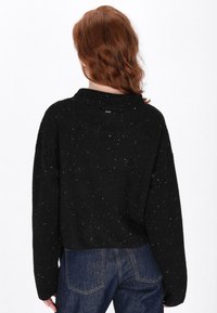 Zwart cropped sweater met een textuurpatroon en witte spikkels. Voorzien van een kraag en een relaxte pasvorm. Gecombineerd met donkerblauwe denim broek.