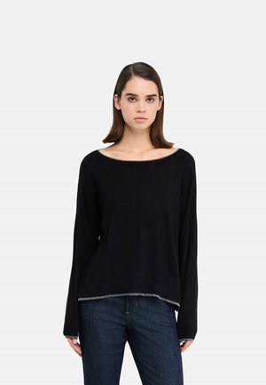 Donna con capelli scuri e lisci, che indossa una maglia nera a maniche lunghe con rifiniture argento e jeans blu, in piedi contro uno sfondo chiaro e uniforme.