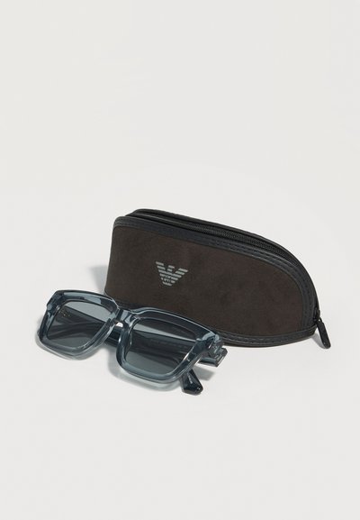 Διάφανα γυαλιά ηλίου με γκρι τετράγωνο πλαίσιο, τοποθετημένα δίπλα σε μια σκούρα θήκη με φερμουάρ και ασημένιο λογότυπο Armani.