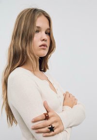 Světlé béžové svetr s žebrovaným vzorem a výstřihem do V, se zapínáním na knoflíky. Na levé ruce jsou dva prsteny: hnědý chlupatý medvěd a černý hvězdicový prsten.