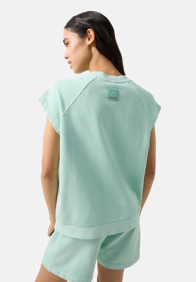 Frau trägt ein mintgrünes, ärmelloses Sweatshirt und passende Shorts, steht mit dem Rücken zur Kamera und zeigt Rückendetails sowie das Logo auf dem Sweatshirt.