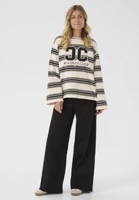 Gestreiftes Langarmoberteil in Creme und Schwarz mit grafischem Logo, kombiniert mit schwarzen weiter geschnittenen Hosen und hellen Schuhen. Glatte Stofftextur.