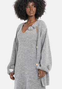 Szary dziergany kardigan z oversize'owymi rękawami i ozdobnymi guzikami w formie pomponów. Posiada dekolt w serek oraz drewniane guziki, wykonany z miękkiego, fakturowanego materiału.