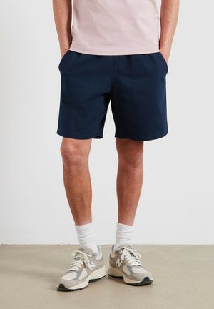 Granatowe spodenki o gładkiej fakturze, z dwoma kieszeniami po bokach i luźnym krojem, zestawione z szaro-białymi tenisówkami oraz białymi skarpetkami.