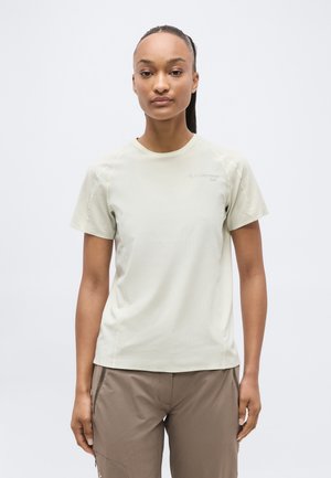 Jeune femme aux longs cheveux bruns attachés en queue de cheval, portant une chemise beige clair à manches courtes et un pantalon taupe, debout devant un fond clair uni.