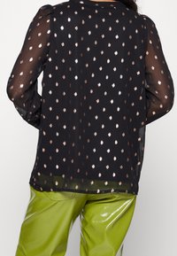 Blouse noire transparente à pois dorés en feuilles, manches longues, épaules discrètes, assortie à un pantalon vert brillant.
