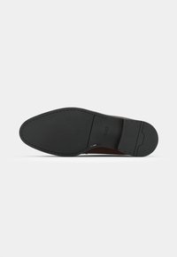 Zapato de cuero marrón con parte superior lisa y suela de goma negra. Presenta detalles de costura a lo largo de los bordes y un diseño minimalista.