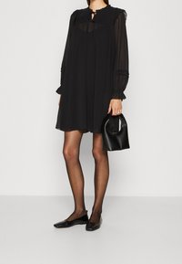 Robe noire à manches longues avec un design plissé, un col texturé, un détail en forme de trou de serrure et des poignets volants. Assortie à un sac à main noir et des ballerines.