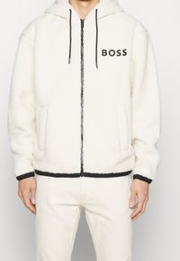 Fleece-Kapuzenjacke in Off-White mit schwarzen Akzenten, Reißverschluss, besticktem "BOSS"-Logo und Fronttaschen. Herrenpassform und Stil.