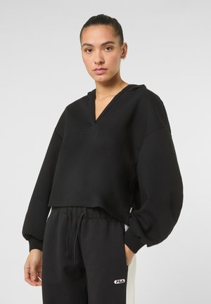 Vrouw met een knot, gekleed in een zwart cropped hoodie en zwarte FILA joggingbroek met witte zijstrepen, tegen een grijze achtergrond.