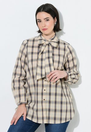 Frau mit kurzen dunklen Haaren, die eine beige-blaue karierte Bluse mit großem Schleifenkragen und blaue Jeans trägt, vor weißem Hintergrund.