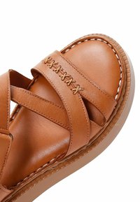 Sandale en cuir marron avec des lanières entrecroisées, des coutures décoratives et une semelle en caoutchouc texturée. Fini lisse et design minimaliste.