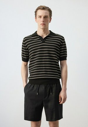 Hombre joven que lleva una camisa tipo polo negra de rayas de manga corta y pantalones cortos negros con cordón, de pie con las manos en los bolsillos sobre un fondo liso.