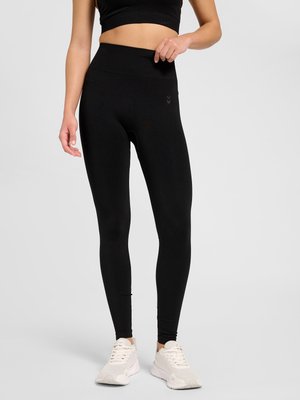 Kvinde iført sorte leggings med høj talje og hvide sneakers, stående mod en ensartet lys baggrund.