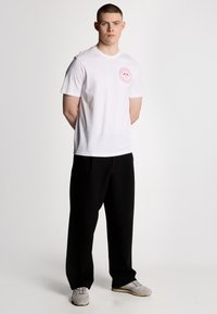 T-shirt bianca in cotone con un logo circolare rosso sul petto, abbinata a pantaloni neri e sneaker grigie, in piedi su uno sfondo neutro.