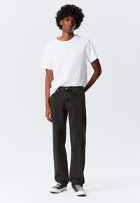 Jeans de ganga pretos com corte direito, combinados com uma t-shirt branca ajustada. O modelo usa ténis pretos e brancos, apresentando um visual casual.