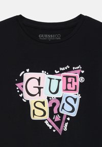Crna majica od organskog pamuka s šarenim blokovnim slovima koja izgledaju kao "GUESS", sa ružičastim trougcckom i rukopisnim naglascima.