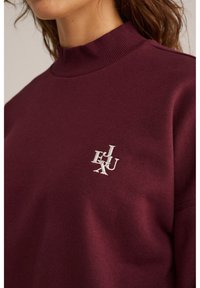 Felpa bordeaux con collo alto realizzata in morbido tessuto, caratterizzata da una finitura testurizzata e un logo bianco ricamato sul lato sinistro del petto.