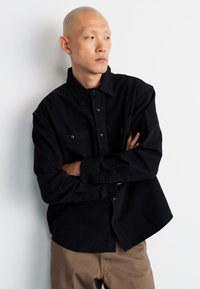 Black button-up shirt with long sleeves, made of soft fabric, featuring two chest pockets and a relaxed fit, paired with beige pants.