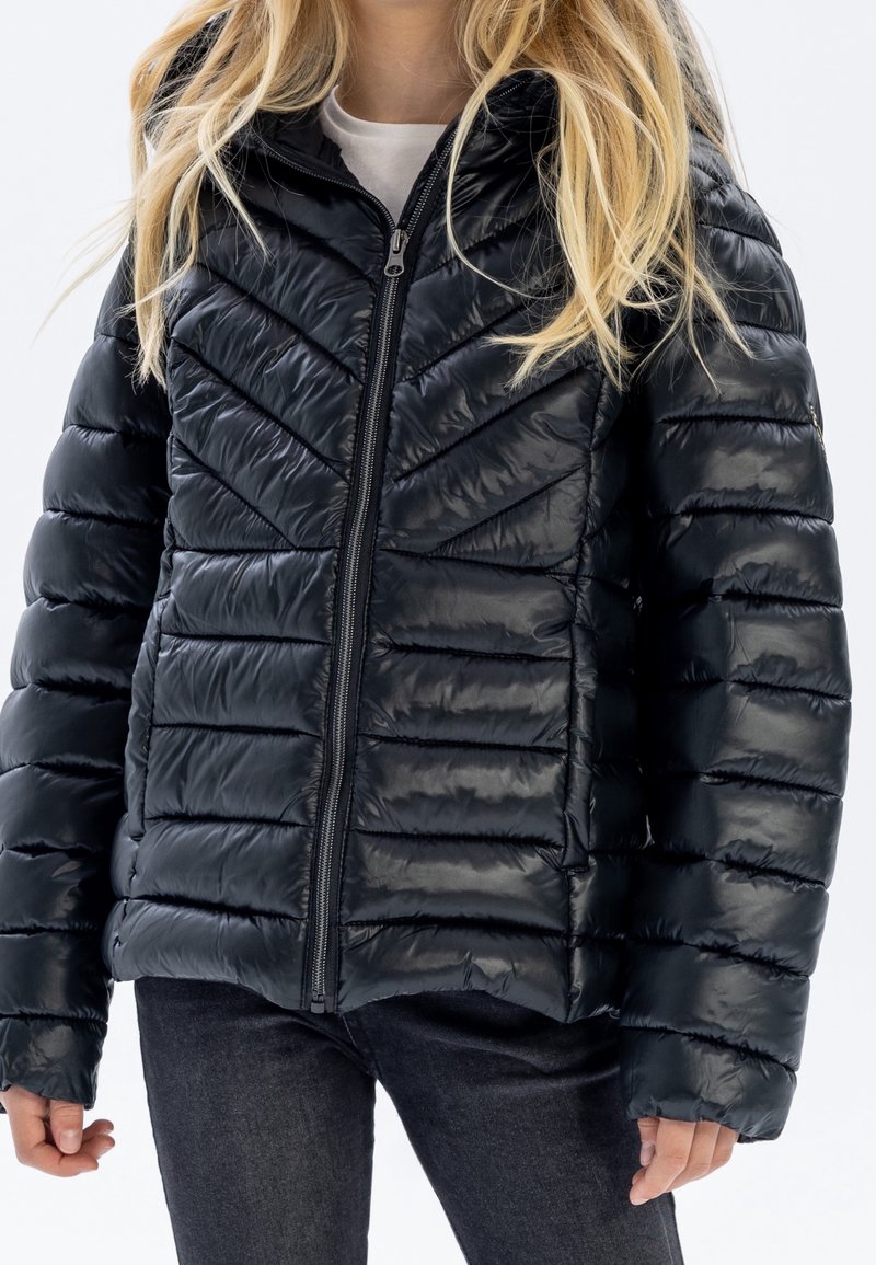 Personne blonde portant une doudoune matelassée noire brillante avec une fermeture éclair, associée à un pantalon sombre sur fond blanc.