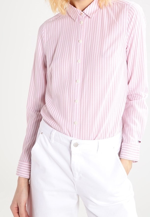 Femme portant une chemise à rayures verticales roses et blanches boutonnée, rentrée dans un pantalon blanc taille haute avec un bouton à l'avant.