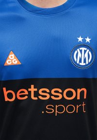 Μπλε και μαύρη αθλητική φανέλα με πορτοκαλί λογότυπο "betsson.sport", πορτοκαλί τρίγωνο λογότυπο "acg" και στρογγυλό έμβλημα με δύο αστέρια.