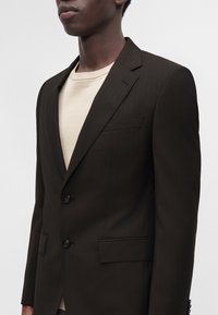 Personne portant un blazer marron foncé sur mesure par-dessus un t-shirt beige clair à col rond, montrée de la poitrine au cou sur un fond uni.