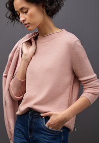 Maglione a maglia rosa chiaro con scollo rotondo, polsini a costine e dettagli laterali, abbinato a jeans blu scuro. Texture liscia e design aderente.