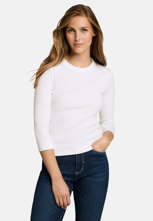 Frau mit langen hellbraunen Haaren, die ein weißes Dreiviertelarm-Top und dunkelblaue Jeans trägt, steht mit einer Hand in der Tasche.